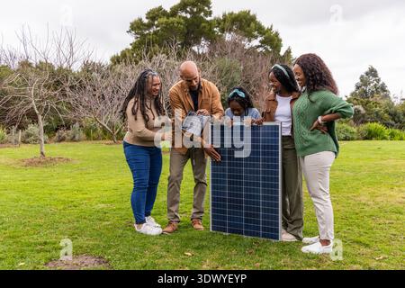 Famiglia afroamericana che impara a conoscere il pannello fotovoltaico su prato erboso, uomo che tiene un piccolo pannello Foto Stock