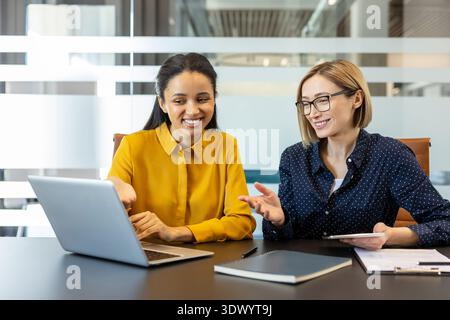 Due donne d'affari multietniche sorridenti che collaborano a un progetto, visualizzano lo schermo di un notebook e discutono idee durante una riunione informale in un ambiente di ufficio moderno Foto Stock