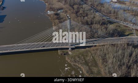 La vista aerea mostra il ponte Swietokrzyski di Varsavia, pilone singolo, cavi bianchi, fiume fangoso, vegetazione invernale sparsa, semaforo su roa a più corsie Foto Stock
