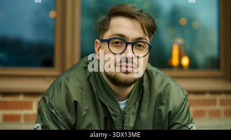 Ritratto ravvicinato di un giovane uomo caucasico barbuto in occhiali rotondi che guarda direttamente la macchina fotografica vicino a un edificio in mattoni. Foto Stock