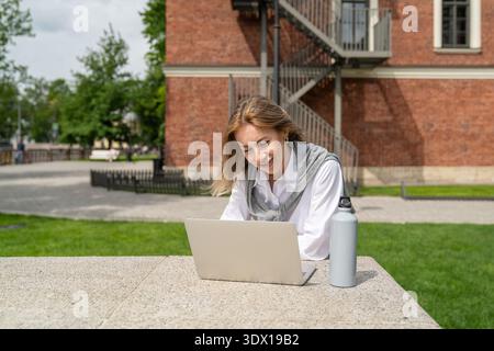 Donna d'affari positiva che lavora a distanza utilizzando un computer portatile all'aperto in un parco, parla tramite videochiamata online Foto Stock