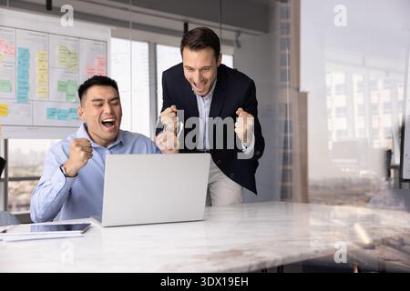 Due uomini d'affari felicissimi urlano di sì pugni d'aria prima dello schermo del notebook Foto Stock