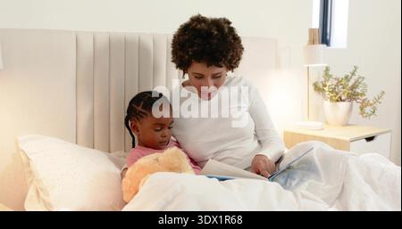 Donna che legge con un bambino seduto nella camera da letto di casa, indossa la parte superiore bianca, tiene un libro aperto e un teddy Foto Stock
