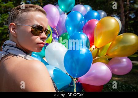 Una giovane donna con i capelli corti e gli occhiali da sole aviatore guarda di lato, posizionata vicino a un grande gruppo di palloncini in lattice lucido in un luminoso ambiente Foto Stock
