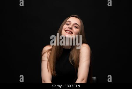 Giovane donna sorridente di sorriso, felice e gioiosa, che posa in studio con sfondo nero, guardando la fotocamera Foto Stock