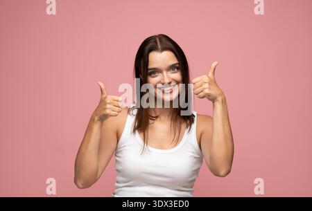 Allegra giovane donna che esprime accordo e ottimismo con un sorriso gioioso e con entrambe le mani che mostrano un gesto con il pollice in alto Foto Stock