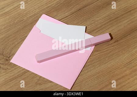 Una busta rosa pastello giace aperta su una superficie di legno, rivelando una carta bianca vuota e un pennarello rosa corrispondente poggiato sulla parte superiore Foto Stock