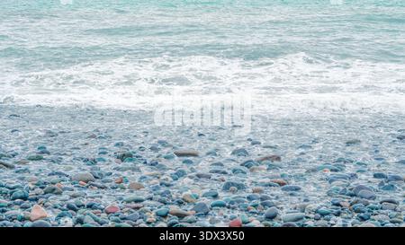 Ciottoli lisci e colorati sparsi su una spiaggia con onde delicate che si lavano sopra di loro Foto Stock