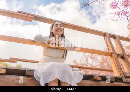 Felice donna asiatica in abito bianco sorridente su un ponte di legno sotto un ciliegio selvaggio dell'himalaya in fiore durante la primavera a Khun Wang, Chiang mai, Thaila Foto Stock