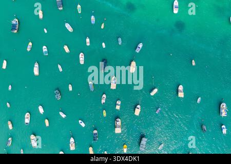 Vista aerea dall'alto verso il basso delle piccole barche ancorate in acque turchesi cristalline al porticciolo di Marsaxlokk, Malta, con punti di ormeggio sparsi e surfa di mare ondulato Foto Stock