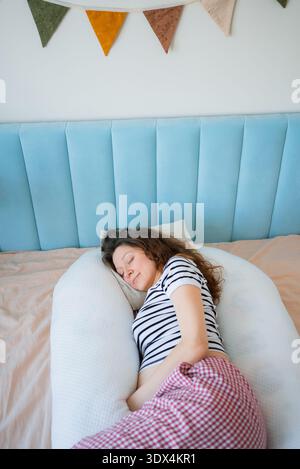 Donna incinta che dorme comodamente al suo fianco con un cuscino per il corpo di maternità di supporto nel letto, promuovendo un sonno prenatale riposante e un delicato sostegno alla pancia durante la gravidanza tardiva Foto Stock
