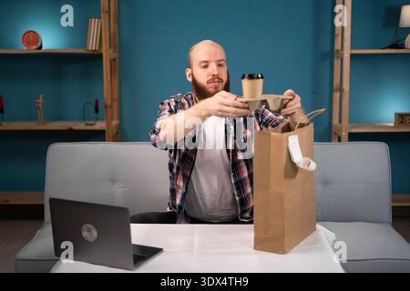 Pausa pranzo uomo barbuto a casa per ordinare e disimballare cibo da asporto e caffè dalla busta di carta, consegna a domicilio. consumo, mangiare e la gente si fa beffe Foto Stock
