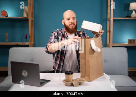 Pausa pranzo uomo barbuto a casa per ordinare e disimballare cibo da asporto e caffè dalla busta di carta, consegna a domicilio. consumo, mangiare e la gente si fa beffe Foto Stock