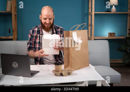 Pausa pranzo uomo barbuto a casa per ordinare e disimballare cibo da asporto e caffè dalla busta di carta, consegna a domicilio. consumo, mangiare e la gente si fa beffe Foto Stock