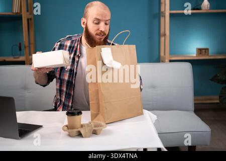 Pausa pranzo uomo barbuto a casa per ordinare e disimballare cibo da asporto e caffè dalla busta di carta, consegna a domicilio. consumo, mangiare e la gente si fa beffe Foto Stock