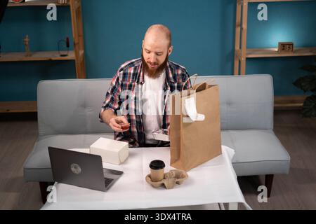 Pausa pranzo uomo barbuto a casa per ordinare e disimballare cibo da asporto e caffè dalla busta di carta, consegna a domicilio. consumo, mangiare e la gente si fa beffe Foto Stock