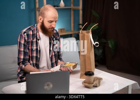 Pausa pranzo uomo barbuto a casa per ordinare e disimballare cibo da asporto e caffè dalla busta di carta, consegna a domicilio. consumo, mangiare e la gente si fa beffe Foto Stock