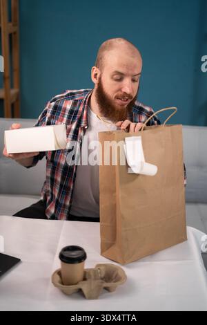 Pausa pranzo uomo barbuto a casa per ordinare e disimballare cibo da asporto e caffè dalla busta di carta, consegna a domicilio. consumo, mangiare e la gente si fa beffe Foto Stock