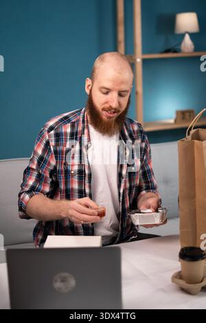 Pausa pranzo uomo barbuto a casa per ordinare e disimballare cibo da asporto e caffè dalla busta di carta, consegna a domicilio. consumo, mangiare e la gente si fa beffe Foto Stock