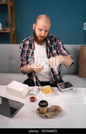 Pausa pranzo uomo barbuto a casa per ordinare e disimballare cibo da asporto e caffè dalla ricevuta di lettura della busta di carta. consumo, consumo, consegna di cibo e.. Foto Stock