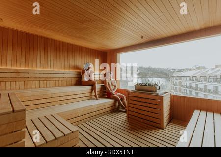 Due persone trascorrono un pomeriggio sereno in una sauna di legno, immergendosi nel calore e nello splendido paesaggio esterno. Foto Stock