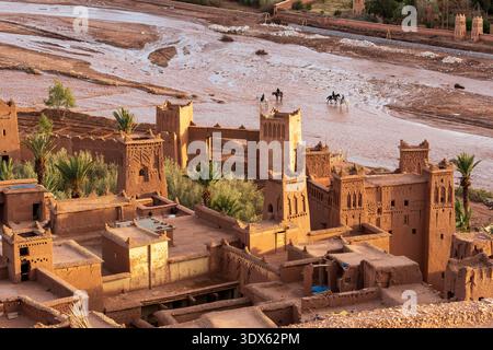 Ait Benhaddou kasbah e cavalieri che attraversano il fiume, montagne dell'alto Atlante, Marocco, 2026 febbraio luce pomeridiana. Foto Stock