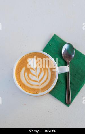 Latte cremoso con delicata latte art a foglia presentato in una tazza bianca su un tovagliolo verde accanto a un cucchiaio. La minima composizione sopraelevata evoca mattine intime Foto Stock