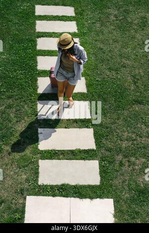 Vista dall'alto di una giovane turista con bagagli che camminano su un sentiero di pietra. Donna viaggiatrice che arriva in hotel o resort durante le vacanze estive Foto Stock