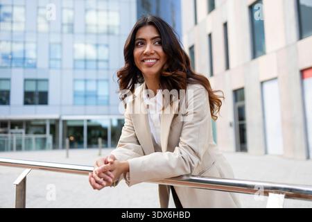 Donna d'affari ispanica in piedi fuori da un moderno edificio di uffici, trasuda fiducia e professionalità. È vestita con un abbigliamento formale, sorridente e.. Foto Stock