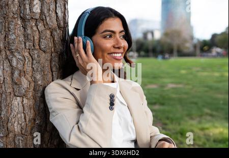 Donna ispanica appoggiata su un albero, indossa cuffie e un blazer beige, sorride mentre ascolta la musica in un parco con un paesaggio urbano sfocato Foto Stock