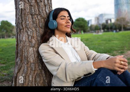 Donna ispanica che indossa le cuffie si siede contro un albero in un parco cittadino, immersa nella musica. Vestita con un abbigliamento da lavoro, gode di un momento di pace in mezzo Foto Stock