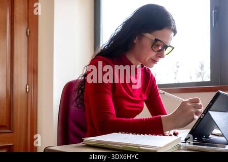 Una donna concentrata che indossa gli occhiali lavora su un tablet digitale con una matita. Si siede accanto a una finestra in una sala ben illuminata, combinando tecnologia e tradizione Foto Stock