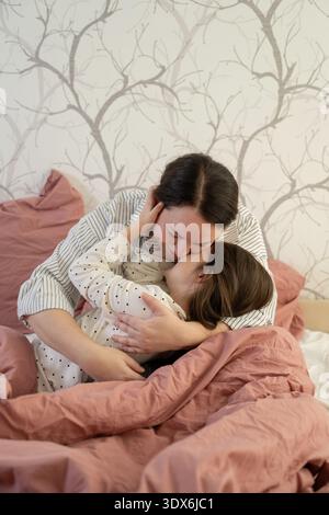 Un momento tenero tra madre e figlia mentre condividono un caldo abbraccio su un letto accogliente. L'ambiente tranquillo della camera da letto arricchisce questa intima famiglia Foto Stock