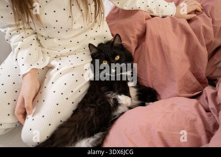 Una ragazza con un abito punteggiato di polka si siede comodamente su un letto, tenendo affettuosamente in mano un gatto bianco e nero rilassato. La scena trasuda calore e tranquillità Foto Stock