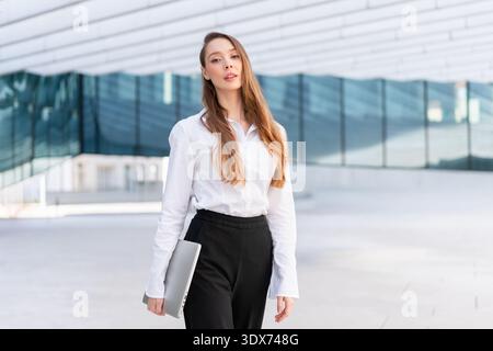 Donna d'affari sicura con camicia bianca e pantaloni neri in piedi con un computer portatile chiuso vicino a un moderno edificio per uffici. Fiducia professionale e concetto di lavoro aziendale Foto Stock