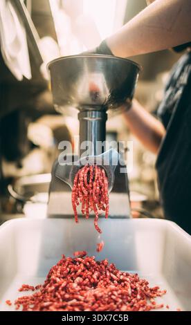 Un primo piano verticale cattura un macinacaffè professionale producendo attivamente carne macinata fresca (macinatura) in un contenitore di plastica bianco. Un guanto h Foto Stock