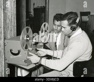 Un uomo che regola un registratore a nastro Magnecord avvolgibile mentre indossa le cuffie, probabilmente ascoltando o modificando audio, Italia anni '1960 Foto Stock