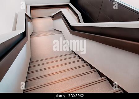 Si affaccia su una scala moderna minimalista con linee geometriche, un corrimano marrone scuro e pareti bianche che creano uno spazio architettonico pulito Foto Stock