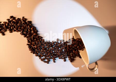 Chicchi di caffè che cadono da una tazza bianca su una superficie leggera Foto Stock
