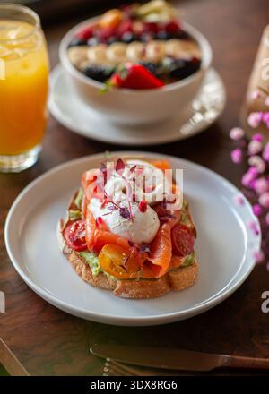 Toast gourmet a lievitazione naturale con avocado schiacciato, strati di salmone affumicato, due uova in camicia e pomodori ciliegini, con un recipiente per frullati di frutta nel Foto Stock