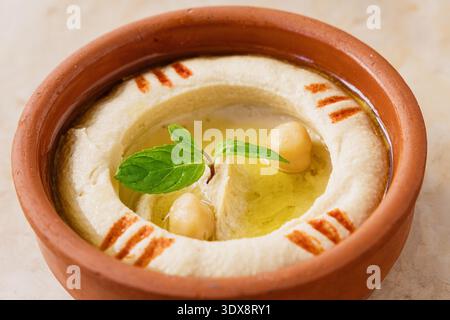 Hummus cremoso servito in una ciotola di argilla rustica, guarnito con ceci interi, menta fresca e un filo di olio d'oliva Foto Stock
