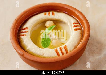 Hummus cremoso servito in una ciotola di argilla rustica, guarnito con ceci interi, menta fresca e un filo di olio d'oliva Foto Stock