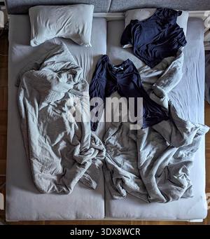 Letto matrimoniale non realizzato con lenzuola sgualcite, due camicie navy spalmate al centro, piumino beige intrecciato, cuscini spinti da parte, luce soffusa sopra la testa, intima Foto Stock
