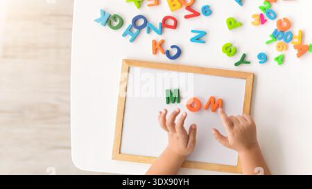 Il bambino esplora le lettere su un tavolo mentre scrive la parola mamma con le lettere in schiuma colorate Foto Stock