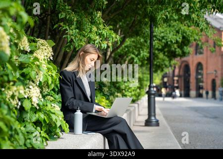 Una donna d'affari sorridente e rilassata lavora con un computer portatile fuori dall'ufficio, ama le giornate estive e le pause giornaliere. Foto Stock