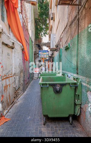 2 dicembre 2025, Bahrein, Manama: Vicolo urbano stretto che mostra i contenitori dei rifiuti verdi lungo un muro testurizzato in una città affollata Foto Stock