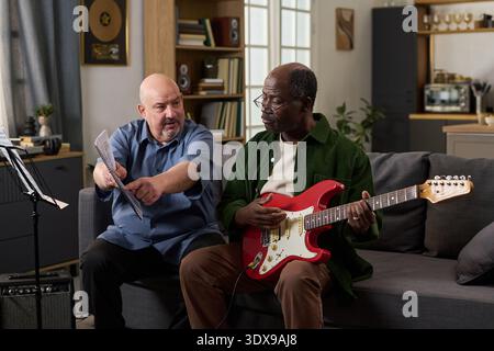 Uomo caucasico di mezza età che mostra spartiti musicali a uomo nero di mezza età che suona la chitarra elettrica sul divano, entrambi seduti nel moderno soggiorno, impegnati in una pratica musicale collaborativa Foto Stock