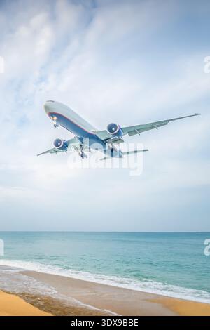 Un aereo vola sopra una spiaggia sabbiosa mentre le onde dell'oceano si schiantano dolcemente sotto. L'acqua blu cristallina riflette il cielo. Le nuvole distanti fluttuano nel Foto Stock