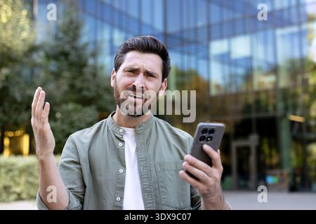 Uomo frustrato che esprime confusione e rabbia mentre incontra problemi di connessione o problemi di app sul suo smartphone, gestendo in modo sprezzante in un ambiente urbano moderno Foto Stock