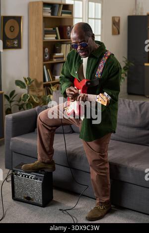 Scatto verticale di Black man anziano che suona la chitarra elettrica mentre si trova in salotto, indossa gli occhiali e si concentra sullo strumento, sull'amplificatore sul pavimento, sugli scaffali e sulle piante sullo sfondo Foto Stock